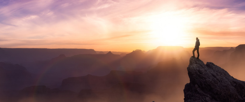 A hiker stands atop a cliff at sunrise overlooking the mountains